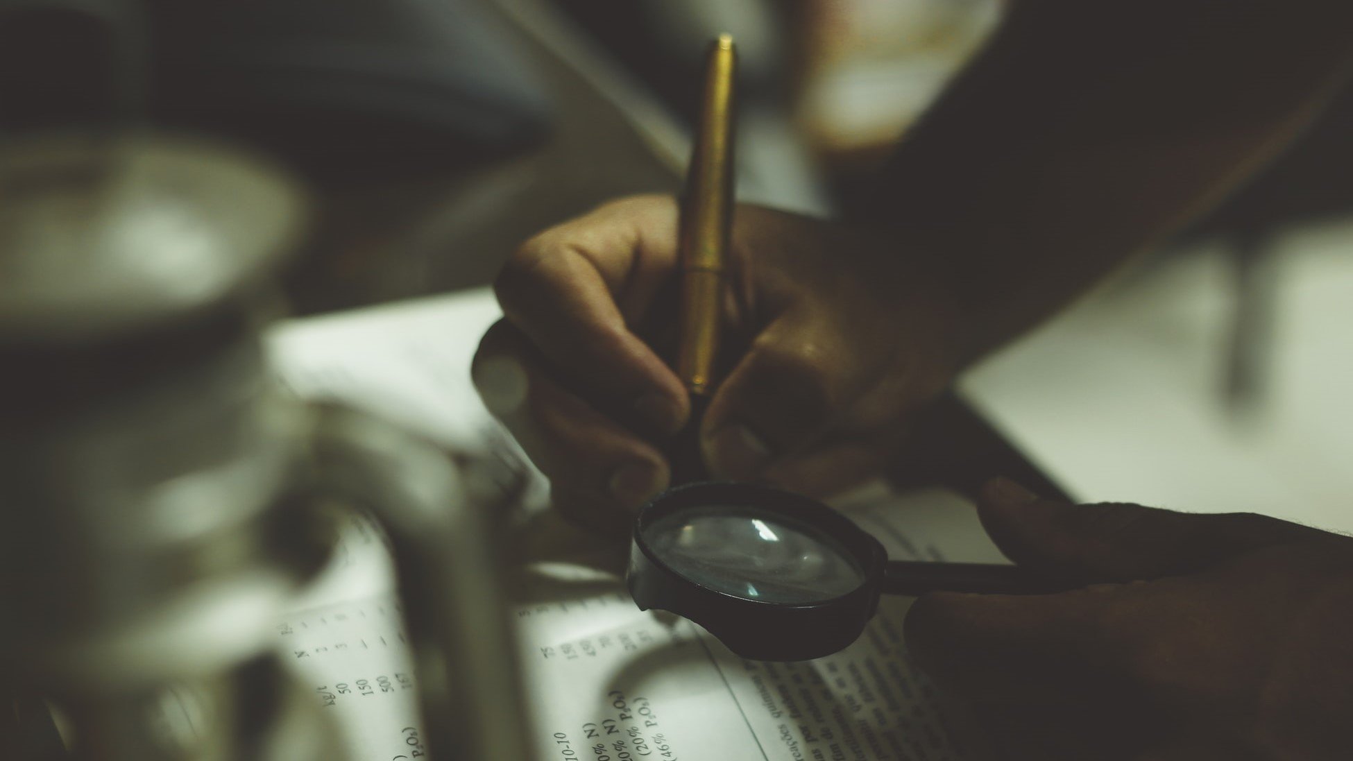 A person examining their ledger book with a magnifying glass and taking notes with a pen.